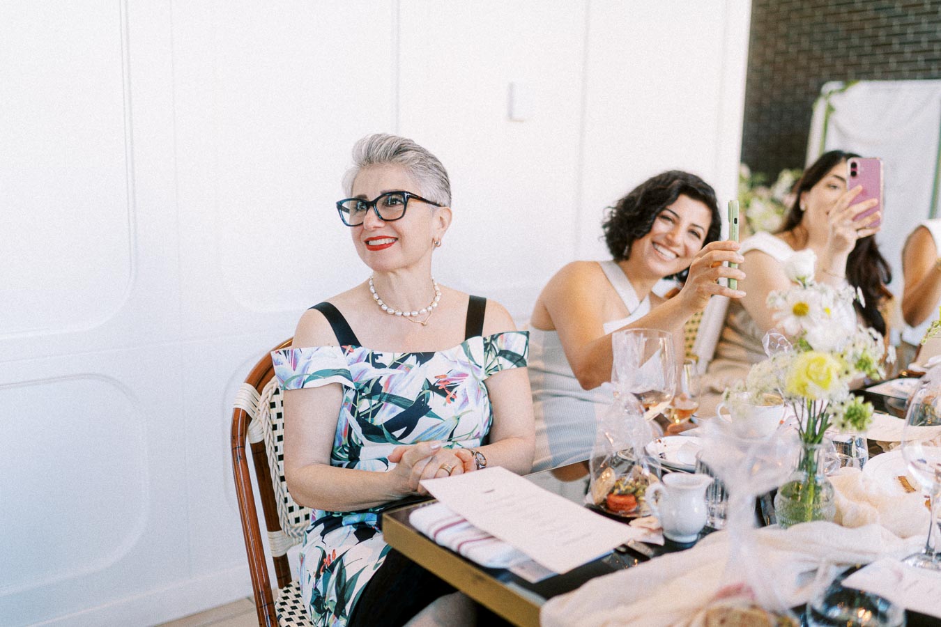 A group of women enjoying a social gathering at a stylish cafe, with one woman in a floral dress smiling and others capturing the moment with their phones, surrounded by beautifully arranged flowers, dishes, and glasses.