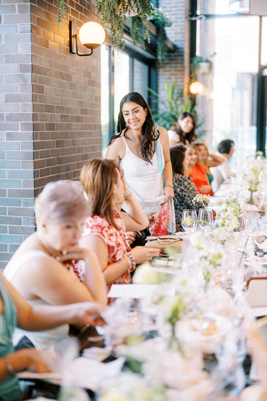 A lively gathering of women enjoying a meal together in a modern restaurant setting, featuring brick walls and hanging plants.
