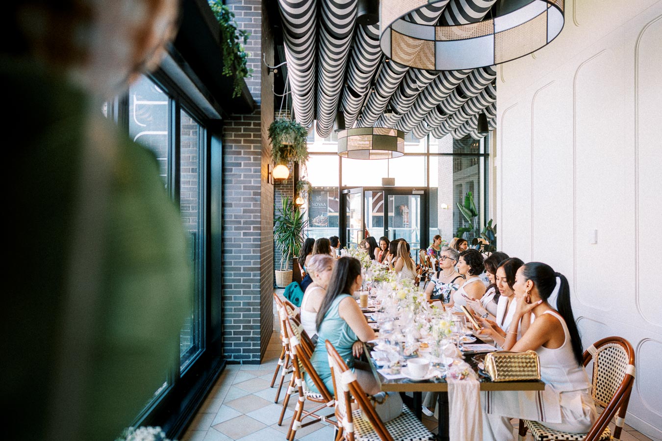 A group of people gather at a long dining table in an elegantly decorated restaurant with large windows and patterned ceiling, enjoying a social event with floral centerpieces.