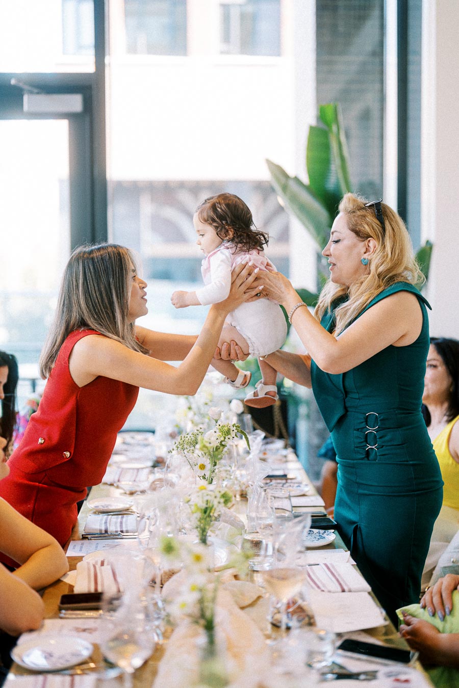 Two women joyfully interacting with a baby at a beautifully set dining table, featuring floral centerpieces and elegant tableware, during a daylight gathering.