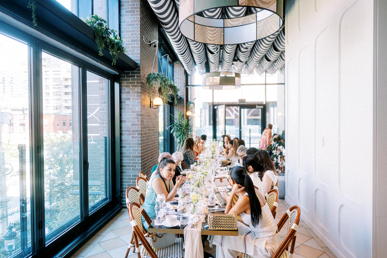 A stylish, modern restaurant interior with a long table set for a gathering. People are seated, engaging in conversation, surrounded by large windows and elegant hanging lights, creating a bright and welcoming atmosphere.