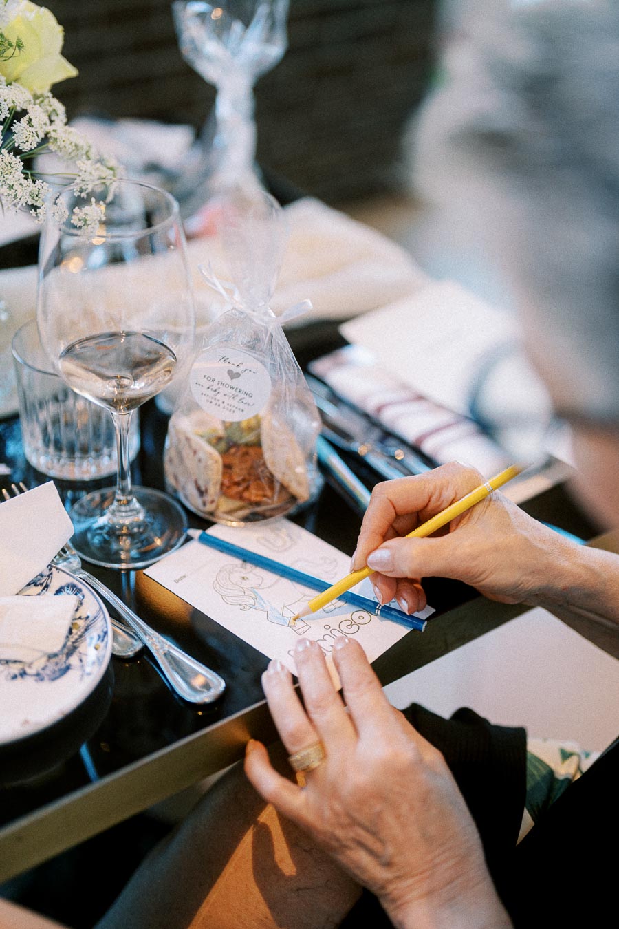 Elderly person coloring a drawing at a restaurant table set with a wine glass, cutlery, and floral decoration. A bag of party favors is visible, enhancing the elegant dining atmosphere.
