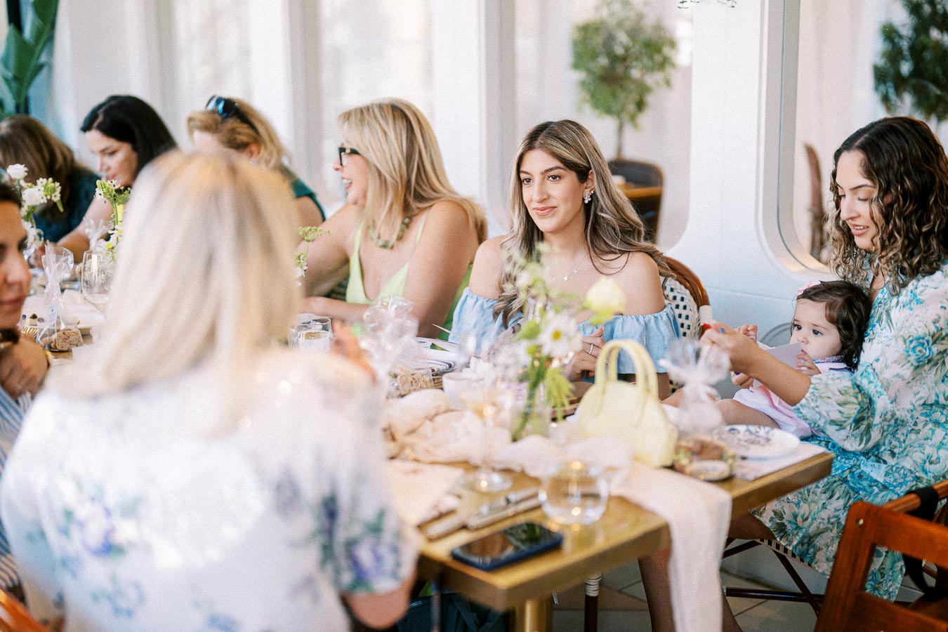 Women engaged in conversation at an elegant luncheon table, adorned with floral arrangements and decorative items, in a brightly lit dining room.