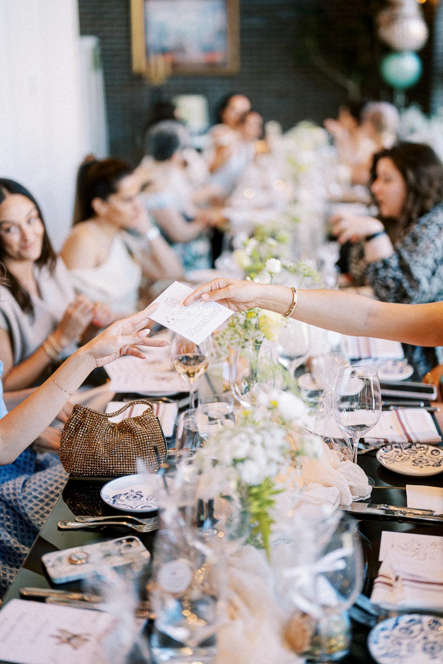 Elegant gathering with people seated at a long table, exchanging cards, amidst a sophisticated setting with floral arrangements and glassware.