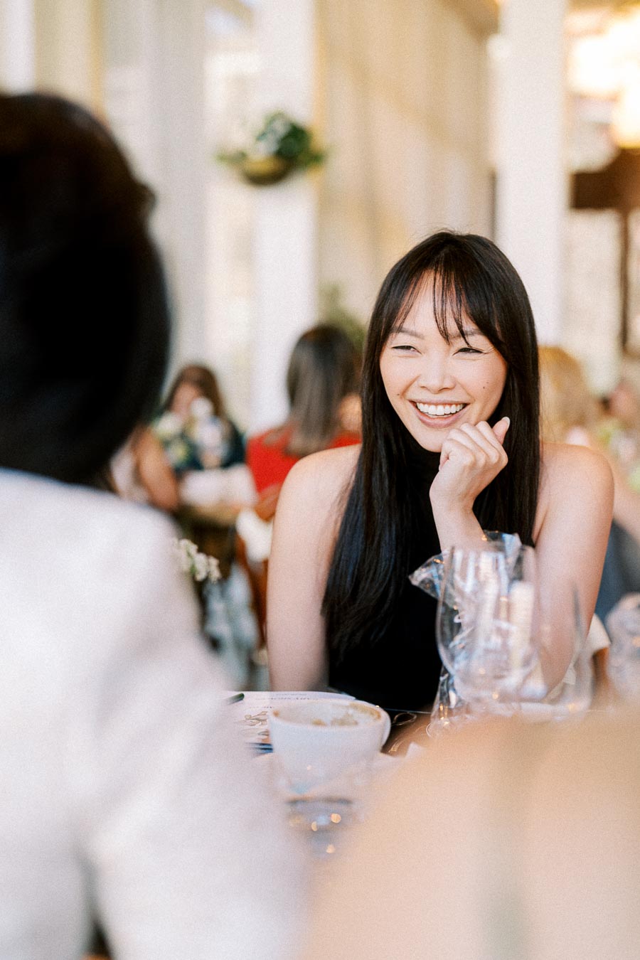 A young woman with long dark hair, smiling and sitting at a restaurant table, surrounded by other diners.