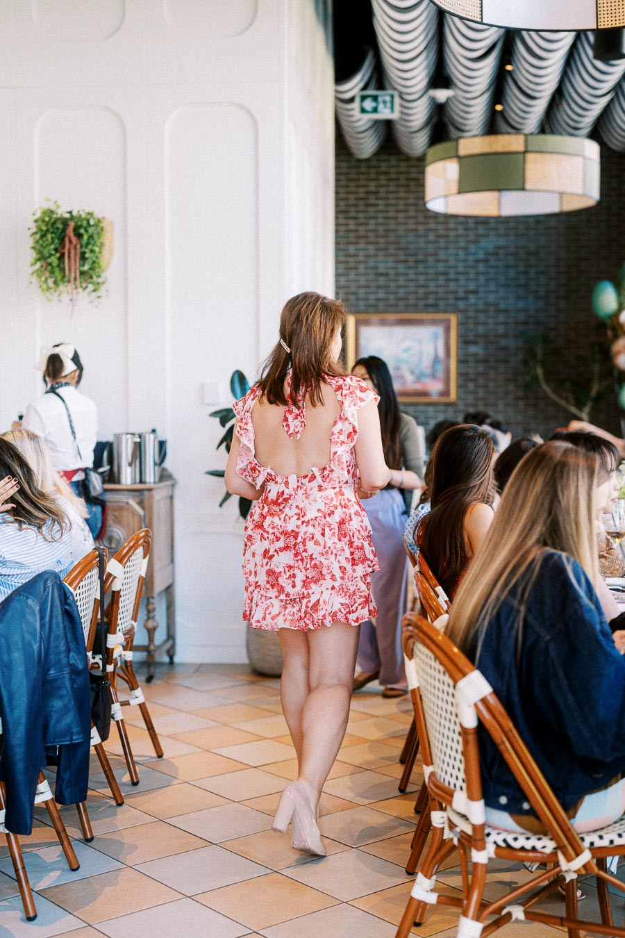 Woman in a floral dress walking through a stylish cafe with patrons seated at tables.