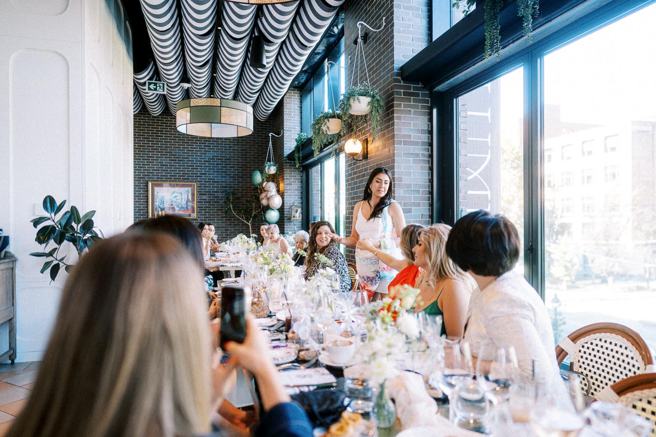 Group of women enjoying a social gathering at a beautifully decorated restaurant, with floral arrangements and elegant table settings. Natural light filters in through large windows, creating a warm and inviting atmosphere.