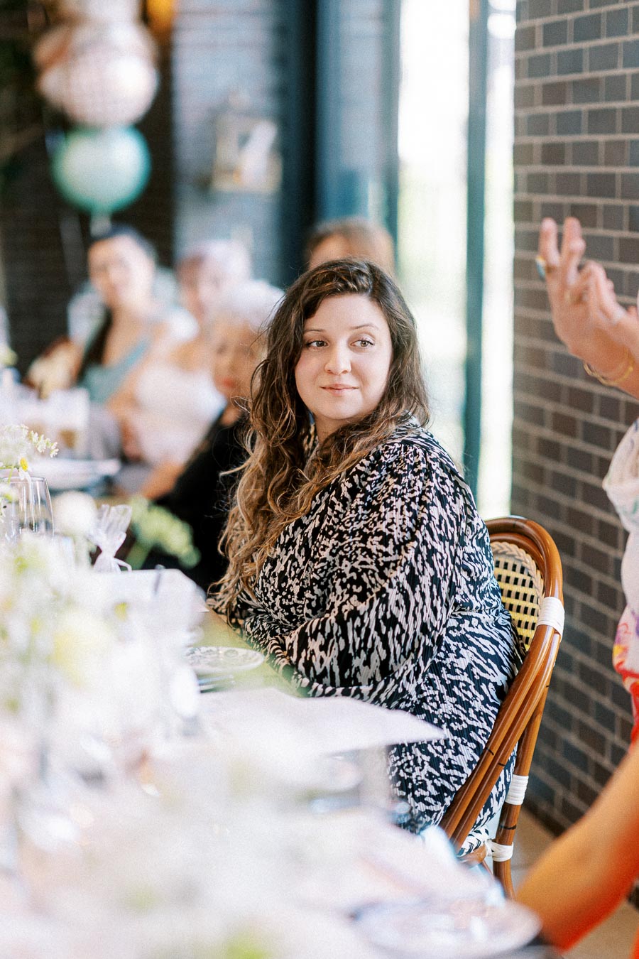 A woman sitting at a dining table during a social gathering, surrounded by blurred guests, with a focus on her thoughtful expression and patterned outfit.