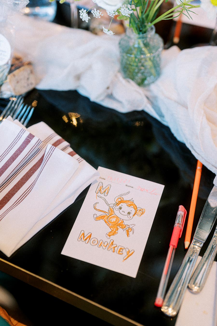 Table setting with a child's drawing of a monkey labeled M Monkey, surrounded by utensils, striped napkins, and a vase with flowers, creating a playful and elegant dining ambiance.