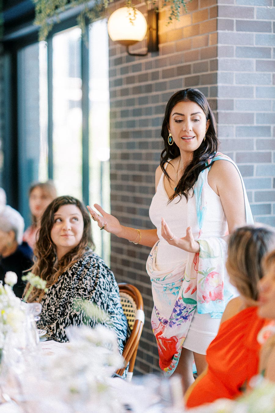 A woman in a white dress and colorful shawl speaks at a gathering in a restaurant setting, with attentive guests seated around a table decorated with flowers.