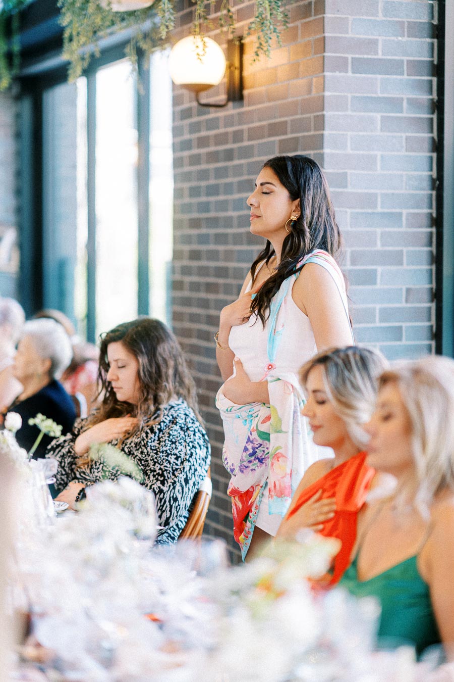 A group of women in bright, colorful dresses and scarves are seated at a decorated table during a gathering, with one woman standing and appearing to be making a heartfelt gesture. The setting features elegant decor and soft lighting, creating a warm and inviting atmosphere.