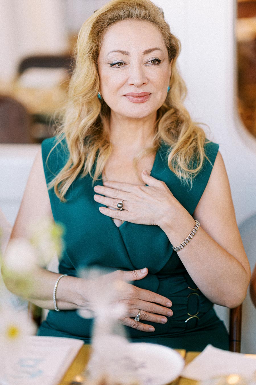 A woman with long blonde hair wearing a teal dress sits at a table, looking content and relaxed. She is holding her hands over her chest, adorned with jewelry, including a ring and a bracelet. The blurred foreground features a table setting with white flowers, suggesting a calm and elegant atmosphere.