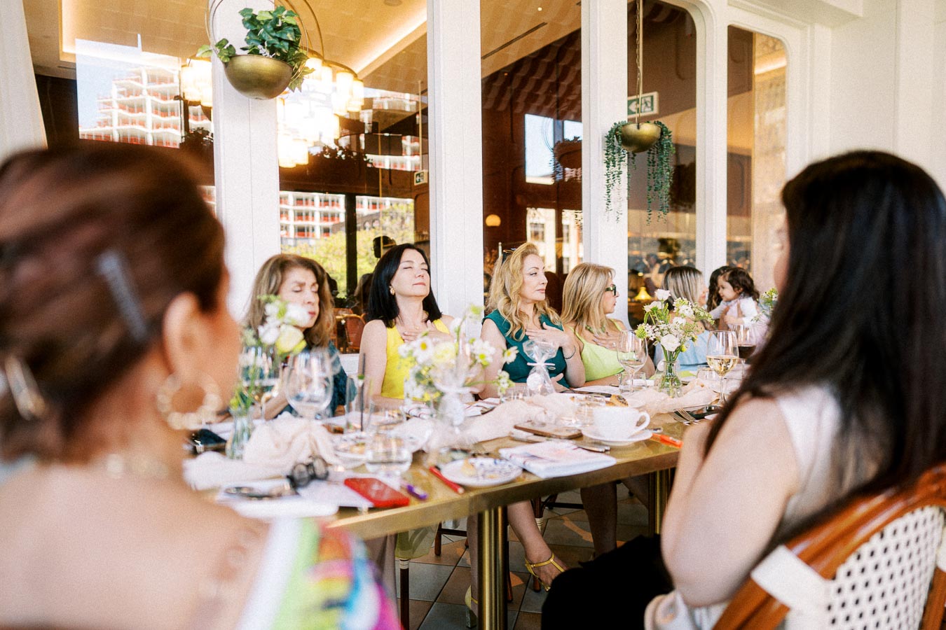 Group of women sitting around a table during a social gathering in a beautifully lit restaurant, with elegant decor and floral centerpieces.