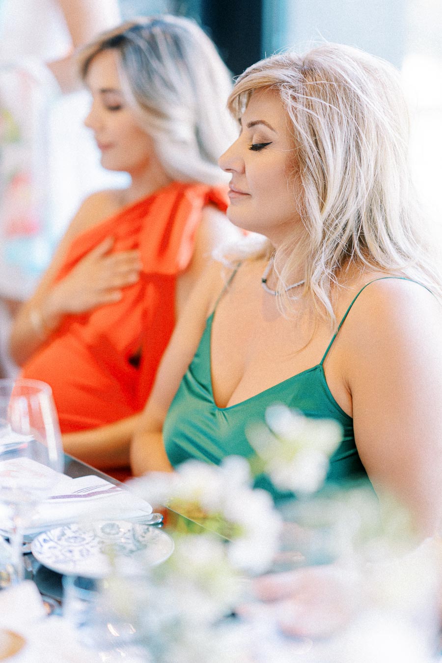 Two women sitting at a table, one in a green dress and the other in red, both with blonde hair, dining in an elegant setting with blurred flowers in the foreground.