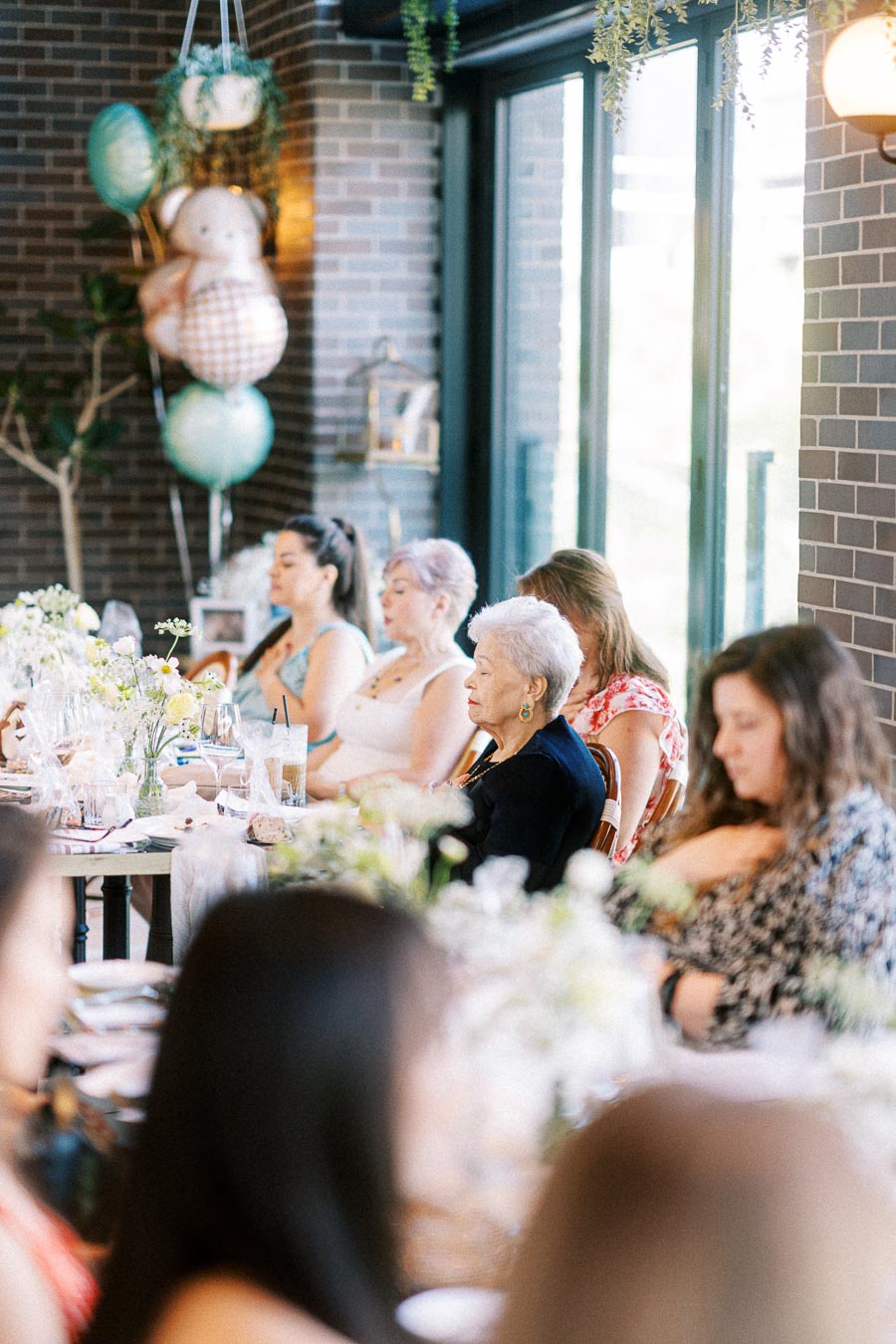 Group of women seated at a long table decorated with flowers and drinks, attending a celebratory gathering in a bright, modern room with brick walls and balloons.