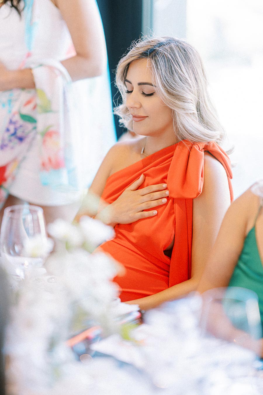 A woman in an elegant orange dress, smiling with her eyes closed, places her hand on her chest at a formal event.