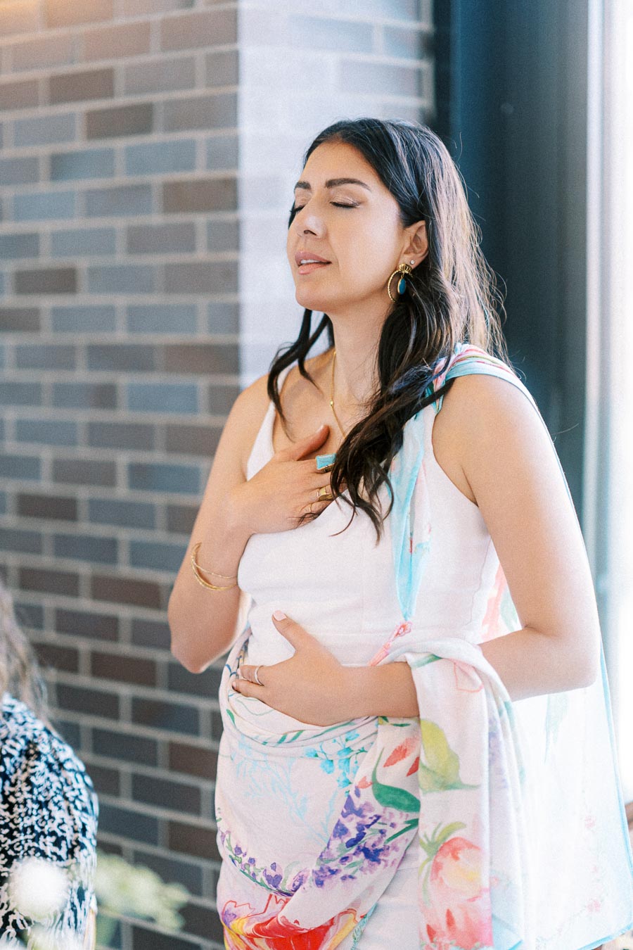 Woman in a colorful shawl meditating near a brick wall, with eyes closed and hands placed on chest and abdomen, in a serene and tranquil setting.