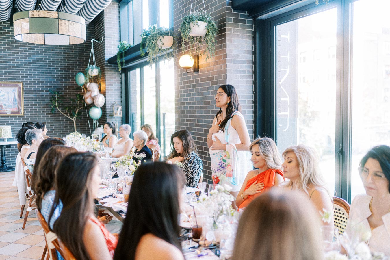 A group of women gathered around a table in a stylish restaurant setting, engaged in a heartfelt conversation. The room features contemporary decor with potted plants and large windows, creating a warm and inviting atmosphere.