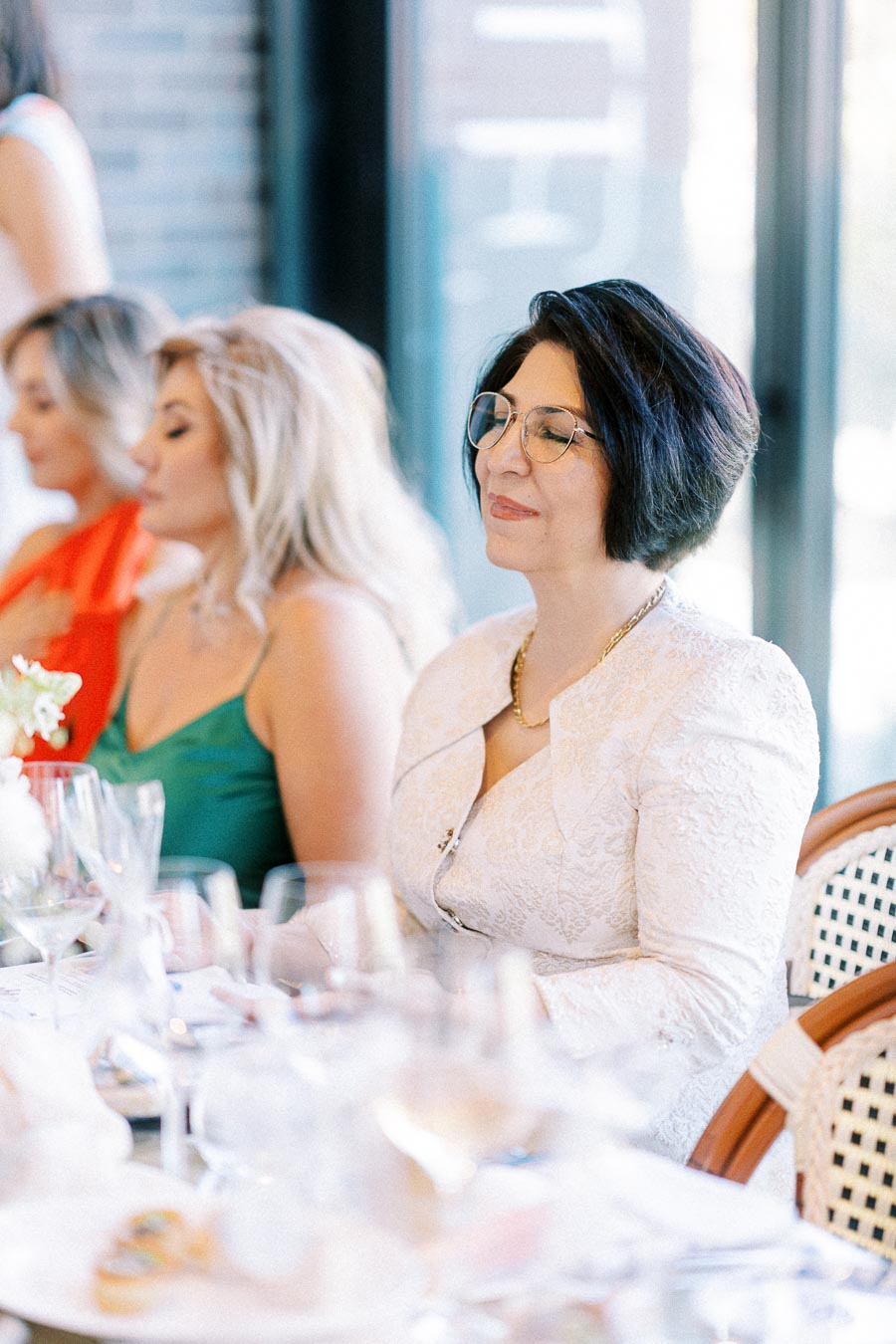 Elegant woman in white lace jacket at formal event, seated at a table with glassware and flowers; guests in background, soft focus.