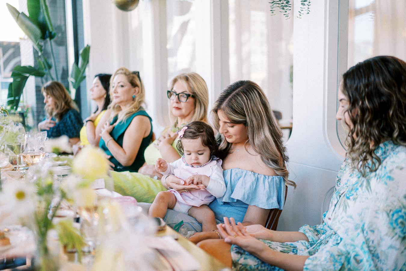 A group of women sitting at a beautifully decorated table during a daytime gathering, with one woman holding a baby and others engaged in conversation.