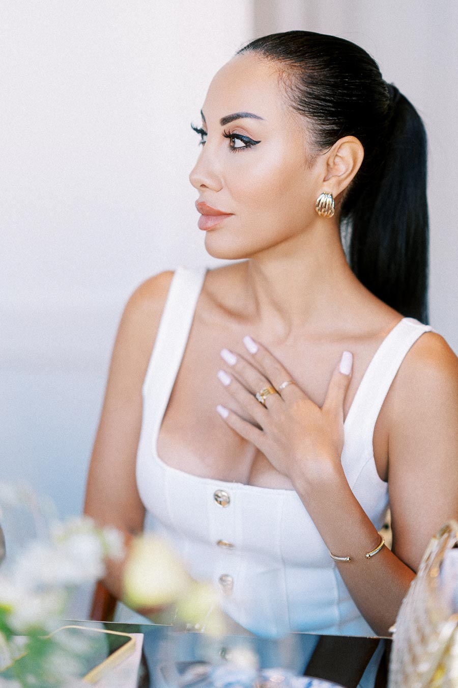 A stylish woman with a sleek ponytail and gold hoop earrings gently places her hand on her chest, showcasing elegant rings while wearing a chic white button-down top.
