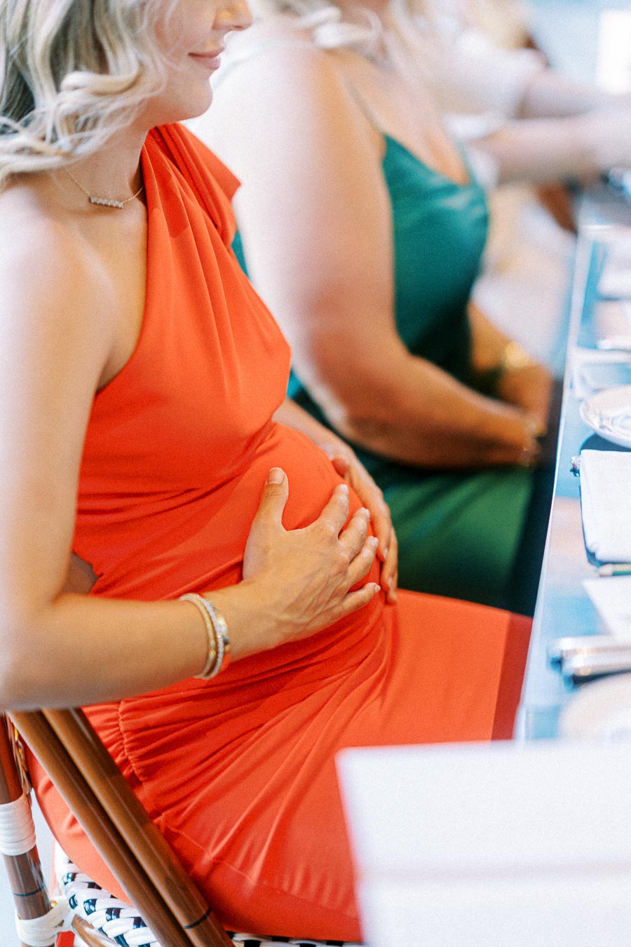 Pregnant woman in an elegant red dress gently holding her baby bump while sitting at a table during a formal event.