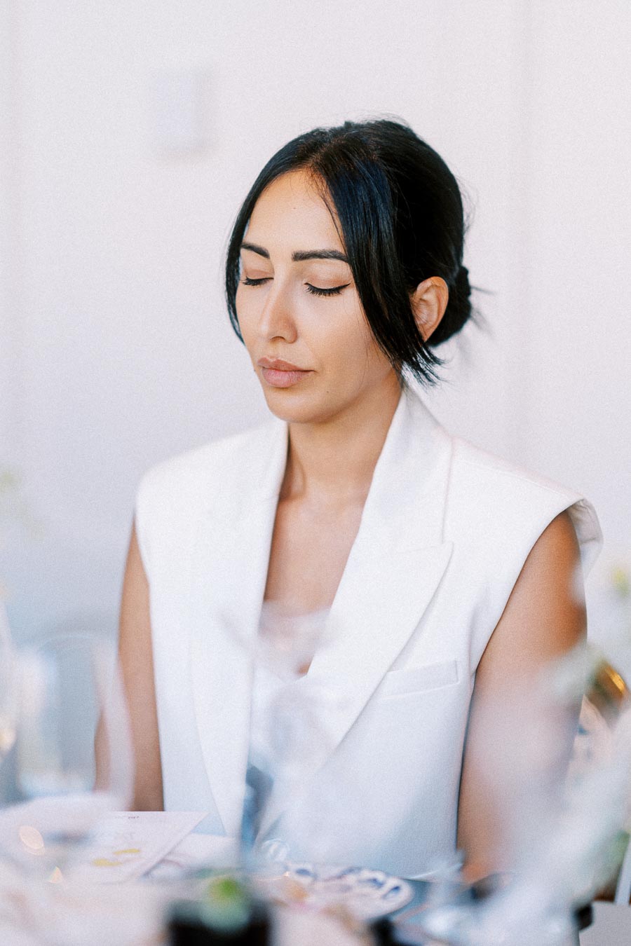 Young woman in a white sleeveless blazer sitting with closed eyes, appearing serene and reflective in a bright indoor setting.