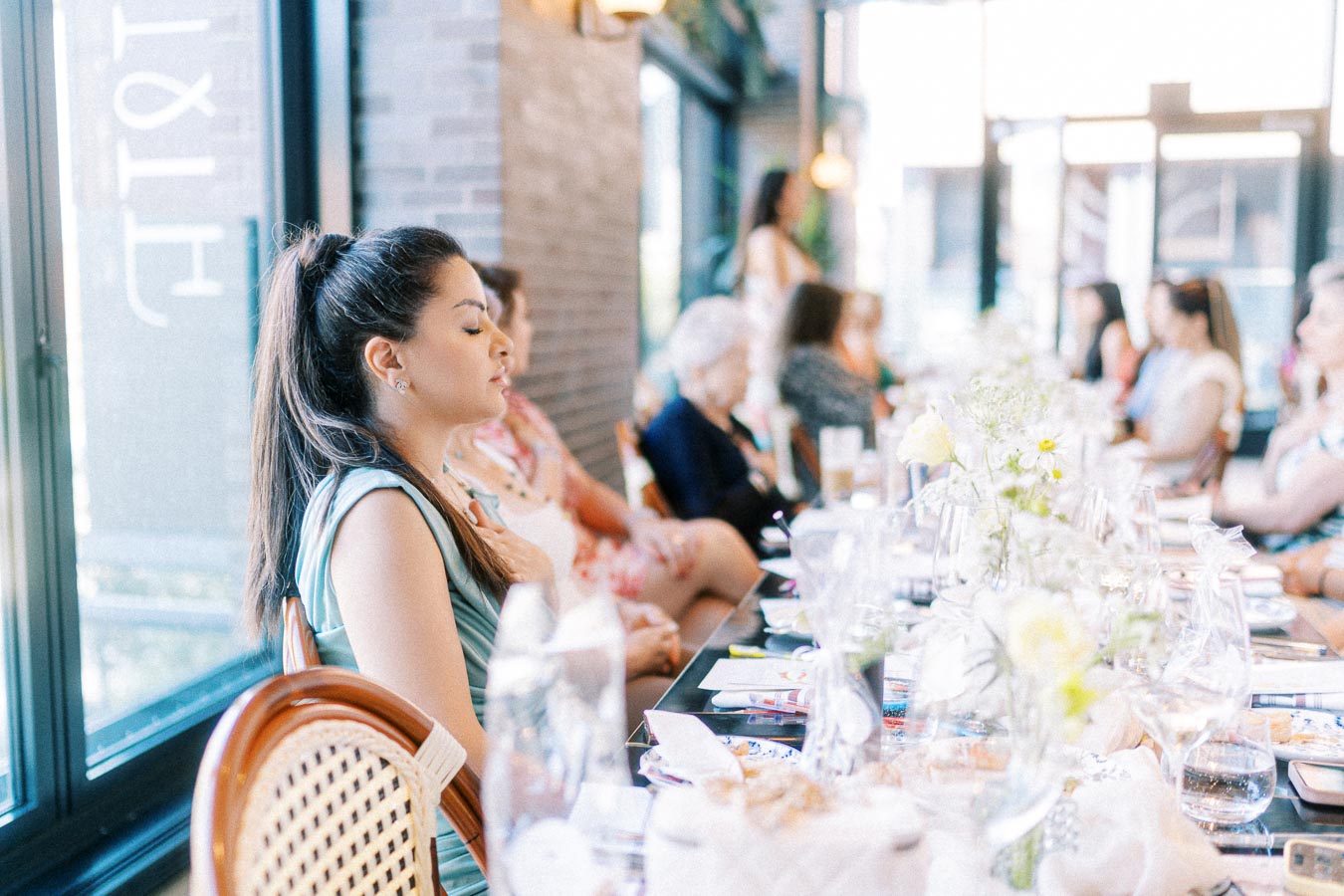 A diverse group of women attentively sit at a beautifully set table during a gathering, surrounded by flowers and natural light from large windows.