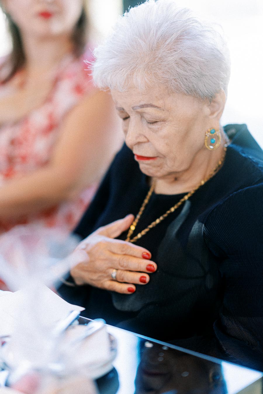 Elderly woman with short white hair and red lipstick sitting with her eyes closed, wearing a navy blue top, earrings, and a necklace, placing her hand on her chest at a table set for a meal.