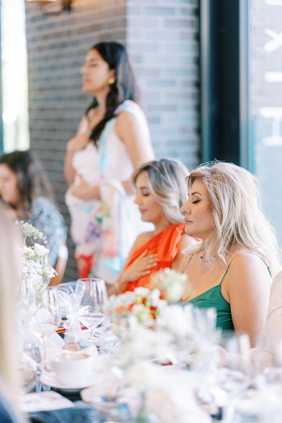 A group of women sitting at a formal luncheon event, engaged in conversation, with elegant table settings and floral decorations in a bright, airy room.