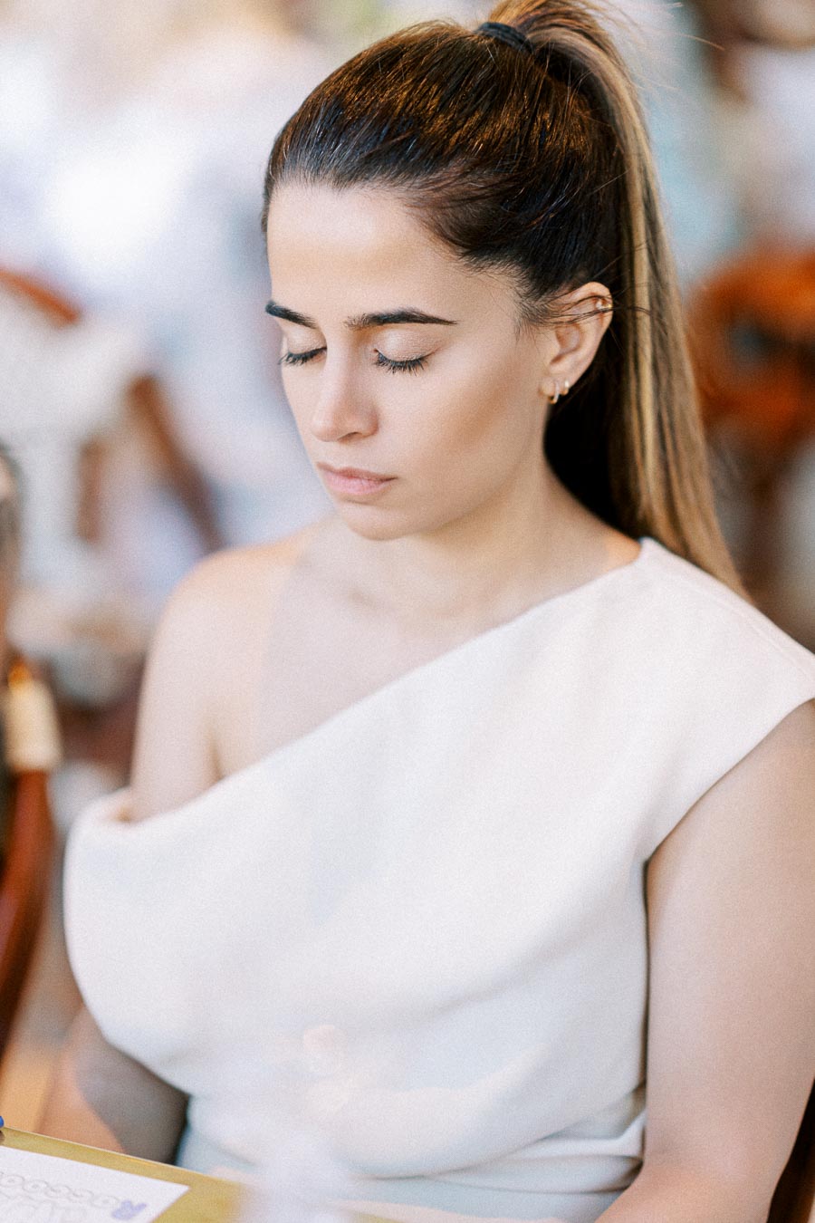 A woman with long hair tied back, wearing a white outfit, sits with eyes closed in a peaceful setting.