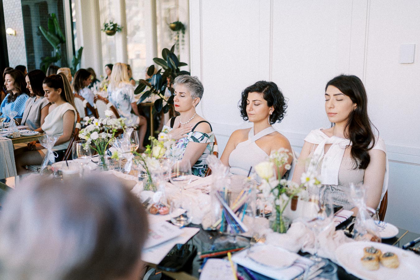 A group of women seated at a beautifully set table in a serene setting, with flowers and elegant decor, engaging in a mindfulness or meditation session.