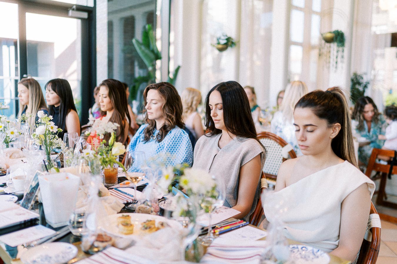 A group of women sitting at an elegantly set dining table, eyes closed, engaging in a mindfulness or meditation session during a daytime gathering, with floral arrangements and natural light in the background.