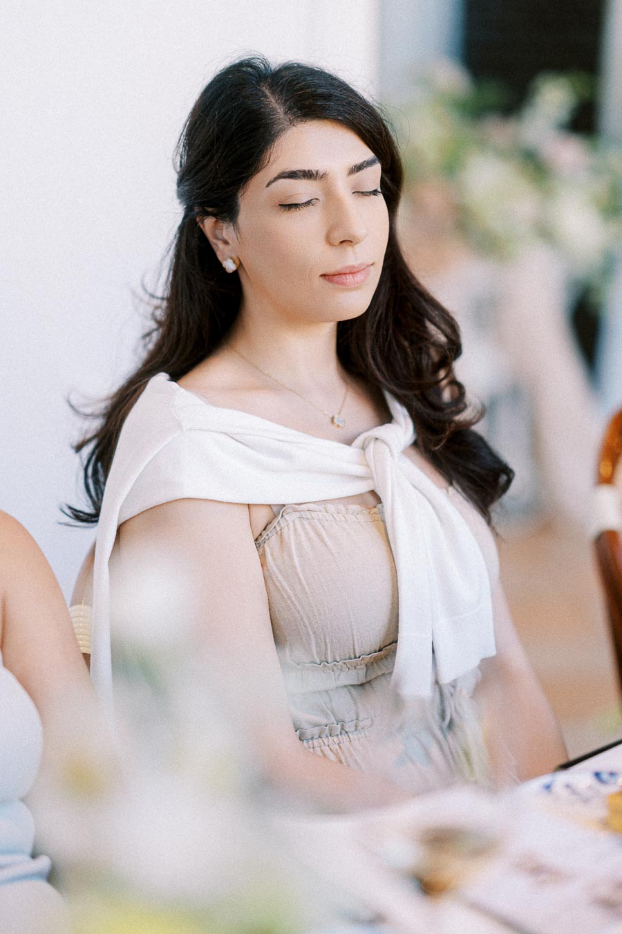A woman with long dark hair wearing a light dress and a white sweater over her shoulders, sitting with eyes closed in a peaceful setting, captured in soft focus with a blurred background.