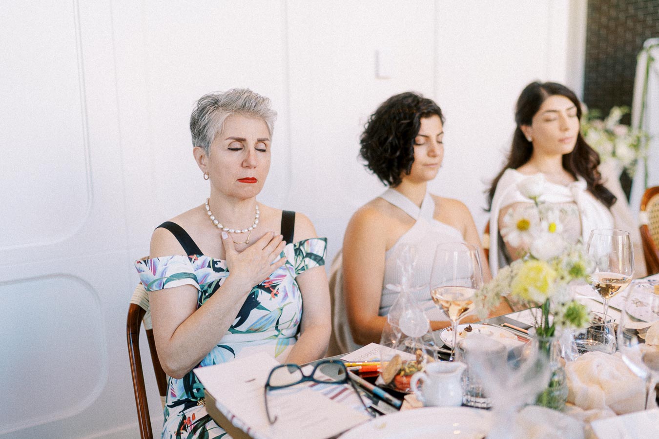 Women practicing mindfulness meditation at a dining table, eyes closed and hands on chest, surrounded by glasses and flower arrangements.