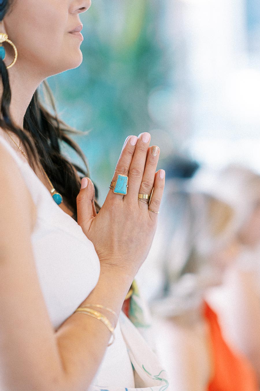 A woman in a white dress holding her hands in a prayer position, wearing a turquoise ring and several gold rings, with a blurred background of soft colors.