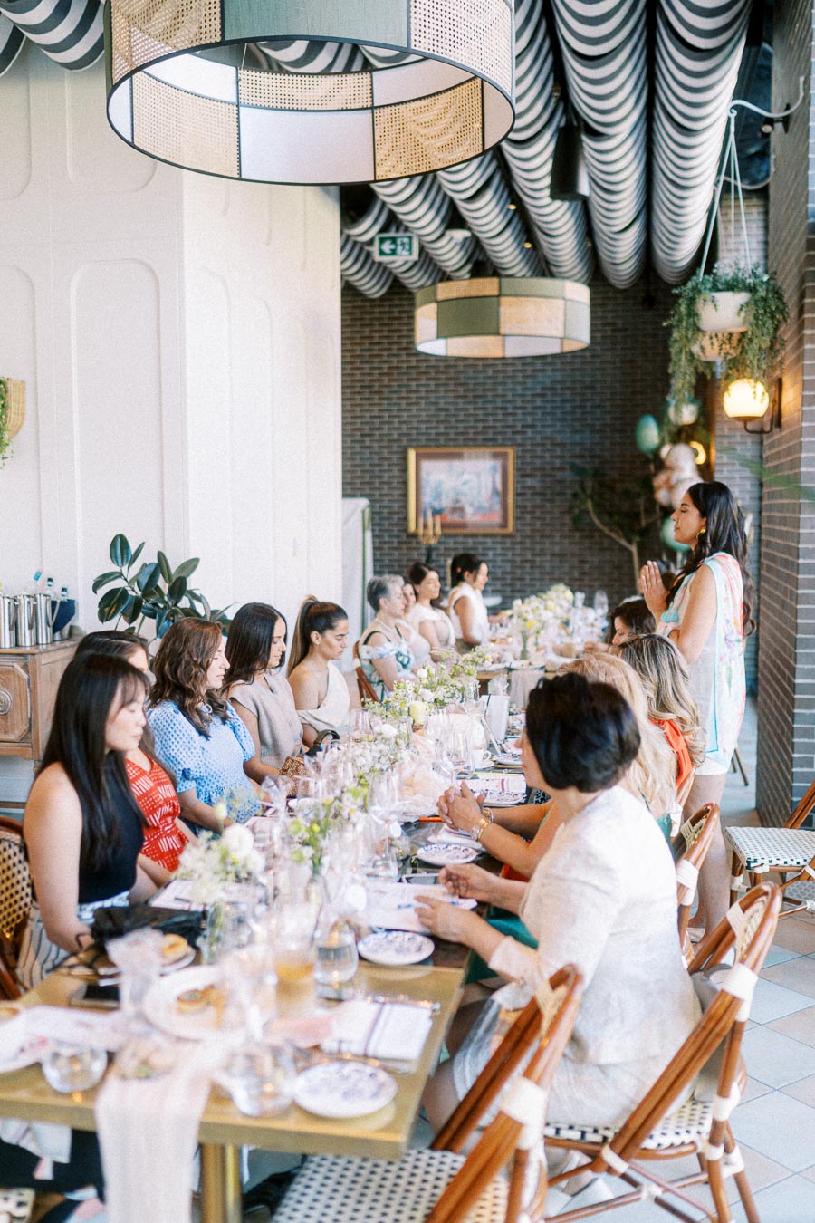 A group of elegantly dressed women seated at a decorated table in a stylish restaurant, engaged in conversation during a daytime gathering.