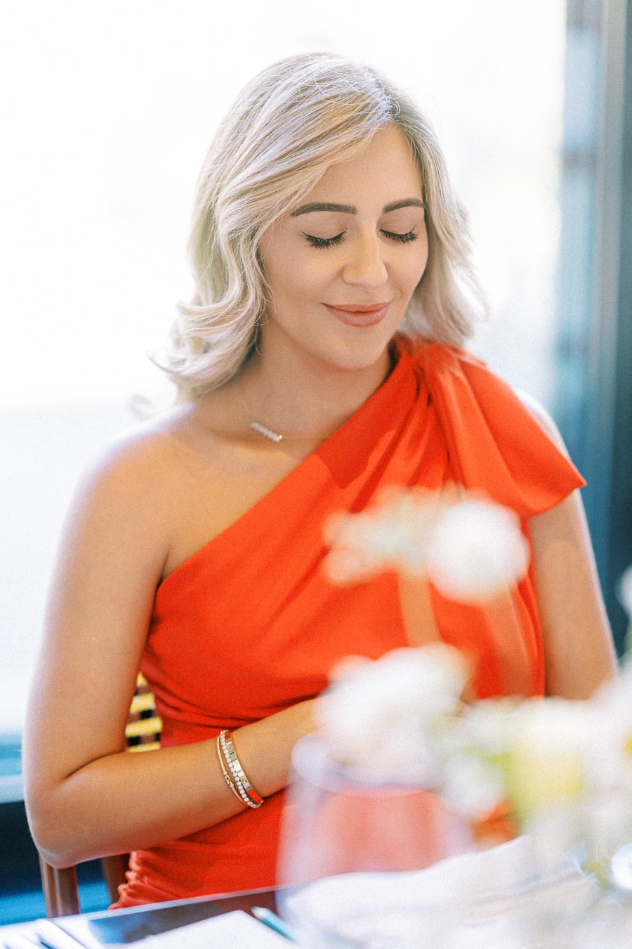 Woman in a vibrant red dress smiling with eyes closed at a dining table, bright atmosphere, elegant setting.