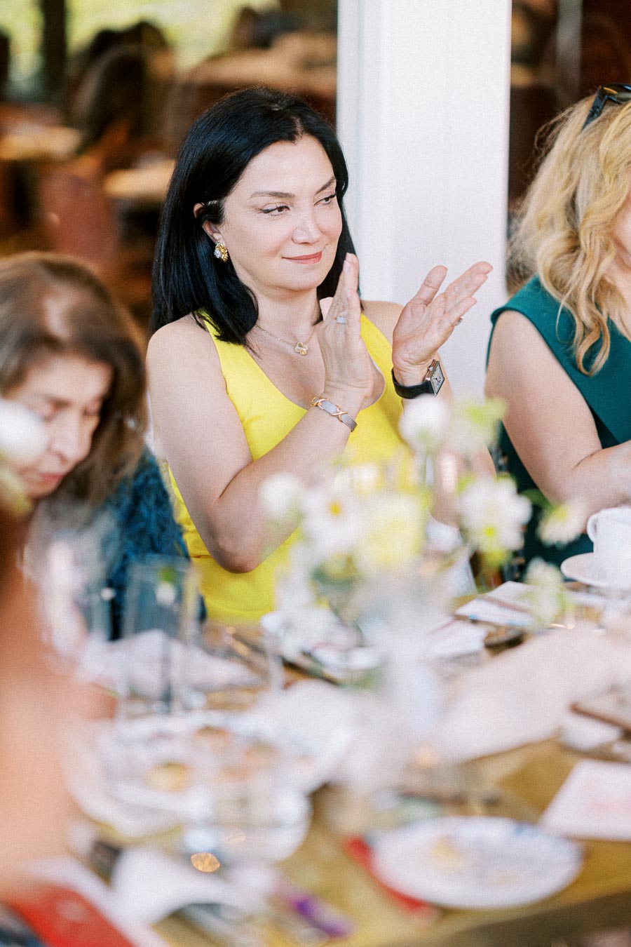 A woman in a yellow dress claps her hands while seated at a dining table decorated with flowers, during a social gathering.