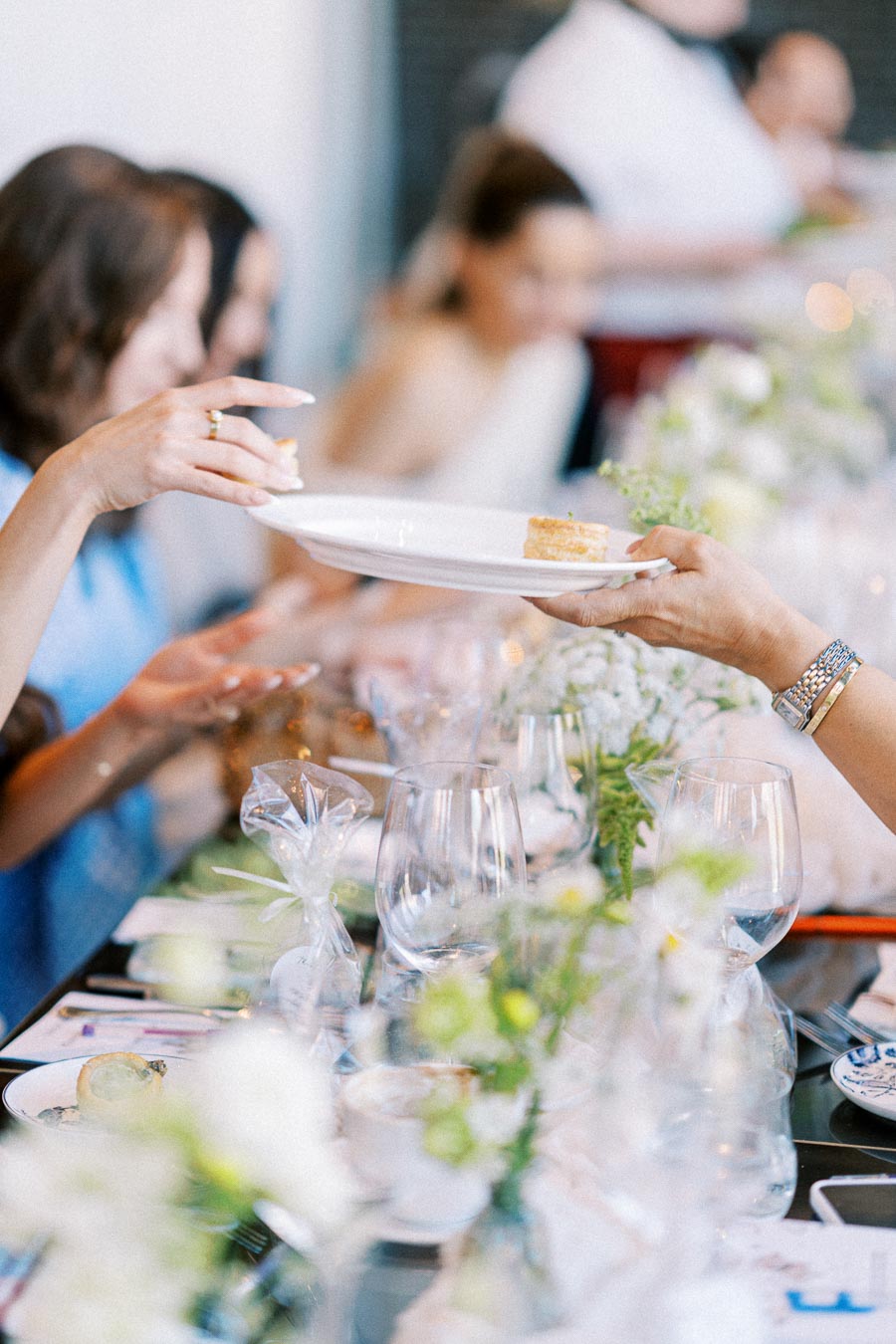 A close-up of a beautifully set dining table during a social event, featuring elegantly arranged floral centerpieces, clear wine glasses, and a person passing a dessert plate with small cakes to another guest.