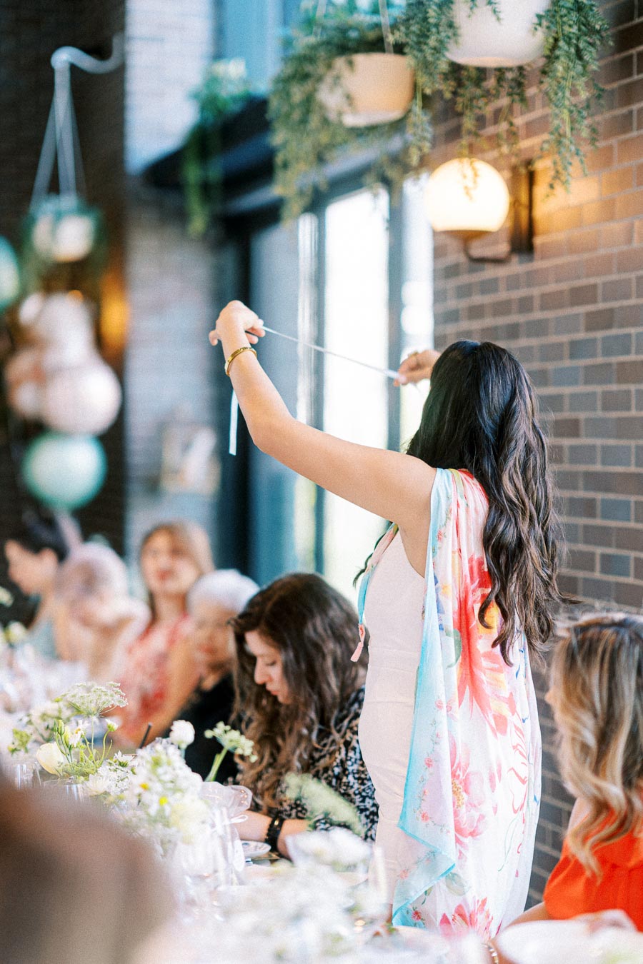 A woman stands at a beautifully set dining table, adjusting a ribbon, with a group of people seated and decorated plants hanging above, creating an elegant and festive atmosphere.