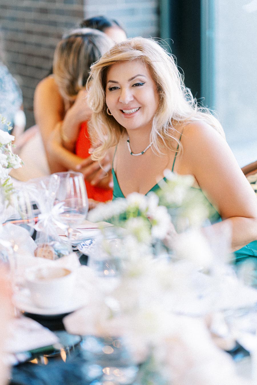 A smiling woman with blonde hair in a green dress sits at a table with elegant tableware, surrounded by blurred floral arrangements at a social gathering.