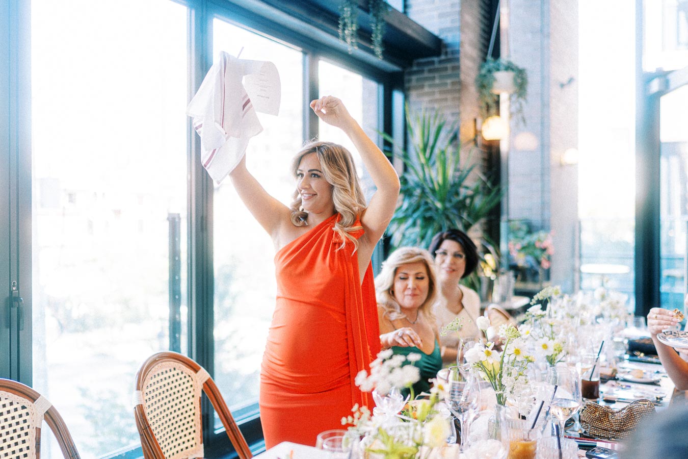 Woman in a vibrant orange dress raises a handkerchief in celebration at a beautifully decorated indoor gathering, with elegant table settings and plants enhancing the ambiance.