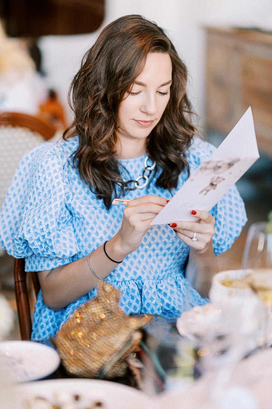 Woman in a blue patterned dress reading a menu at a restaurant, seated at a table with dishes and a glass of wine.