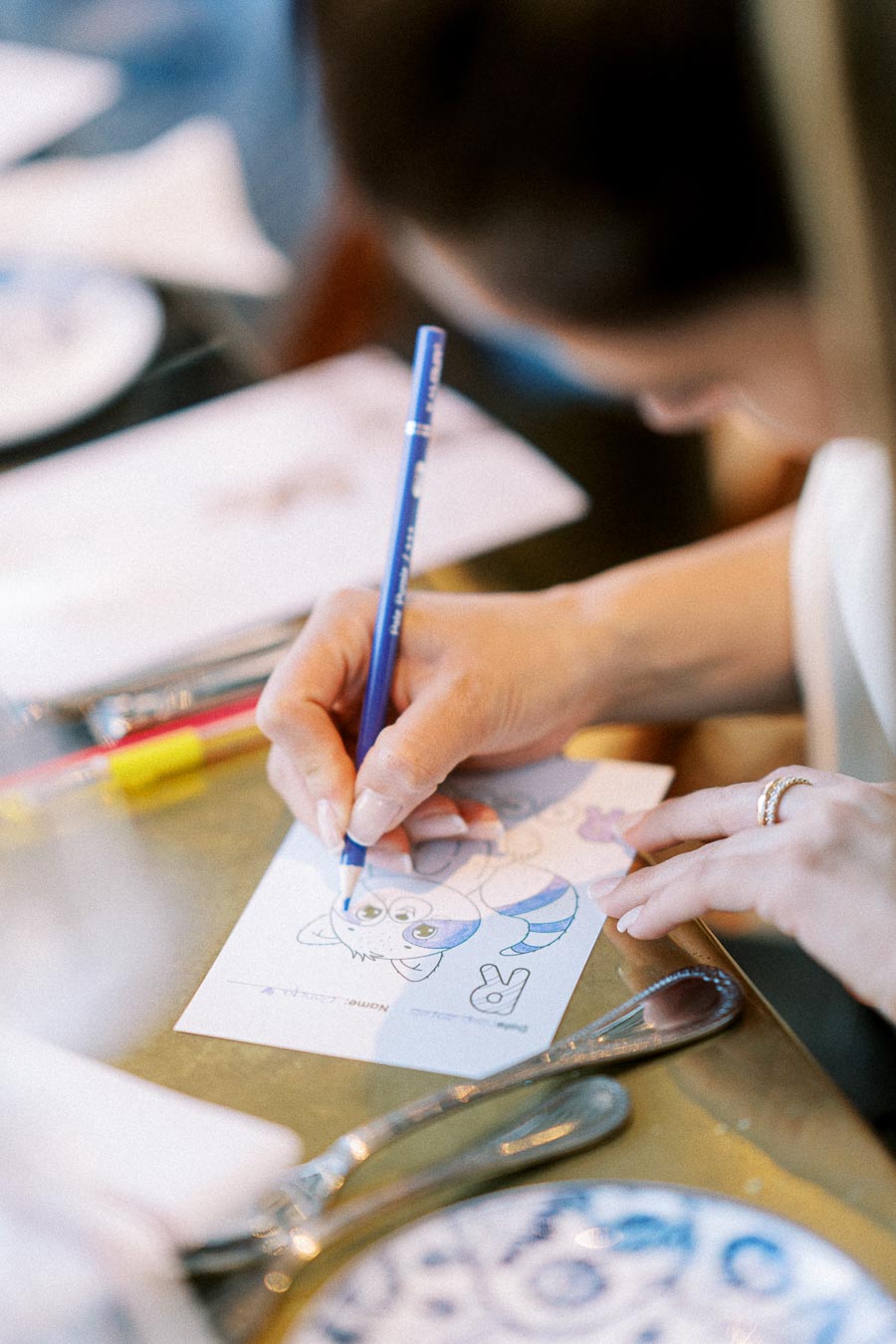 A close-up of a person coloring a cat illustration on a postcard with a blue pencil, surrounded by drawing materials on a table.
