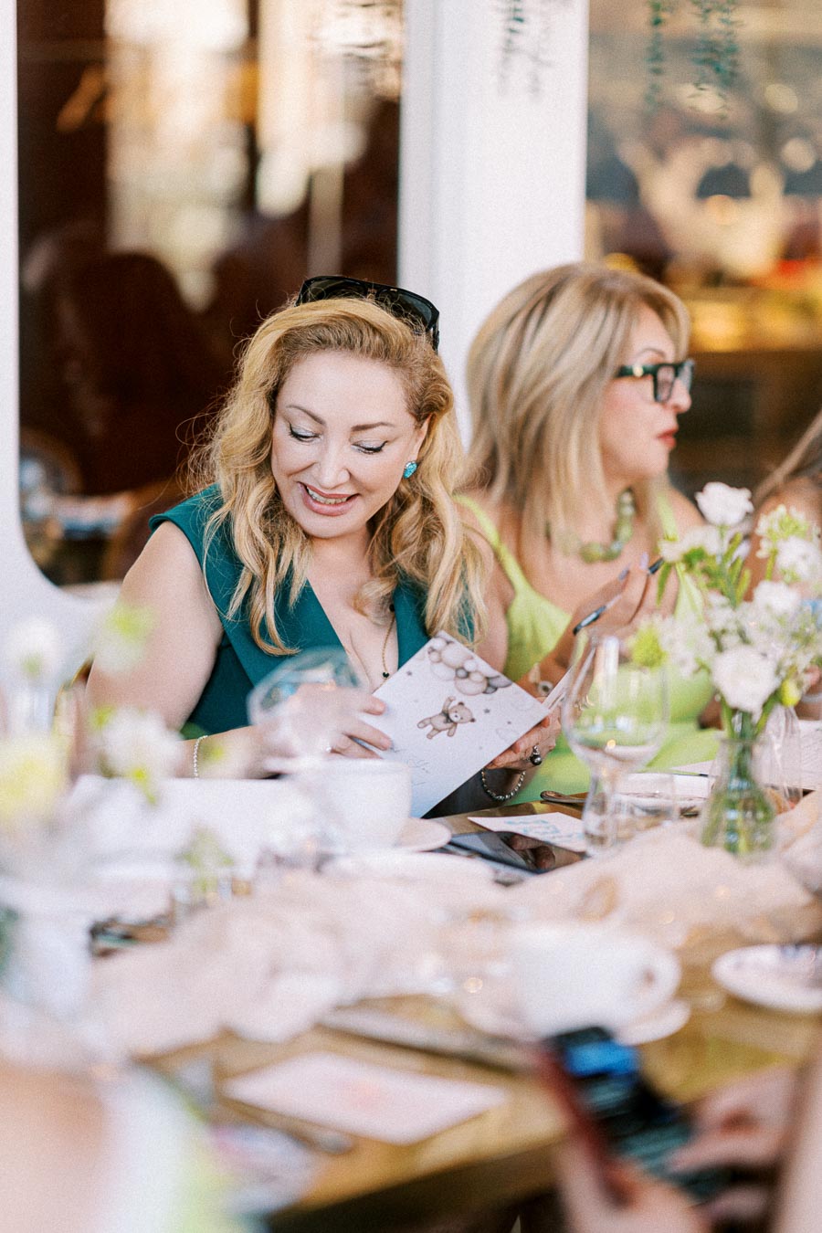Two women enjoying a celebratory gathering at a beautifully set table, one reading a greeting card with a bear design; adorned with flowers and tableware, creating a joyous and elegant atmosphere.
