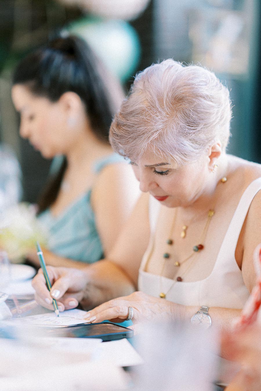 Elderly woman focused on writing at a creative workshop, wearing a white dress and necklace, seated next to another participant.