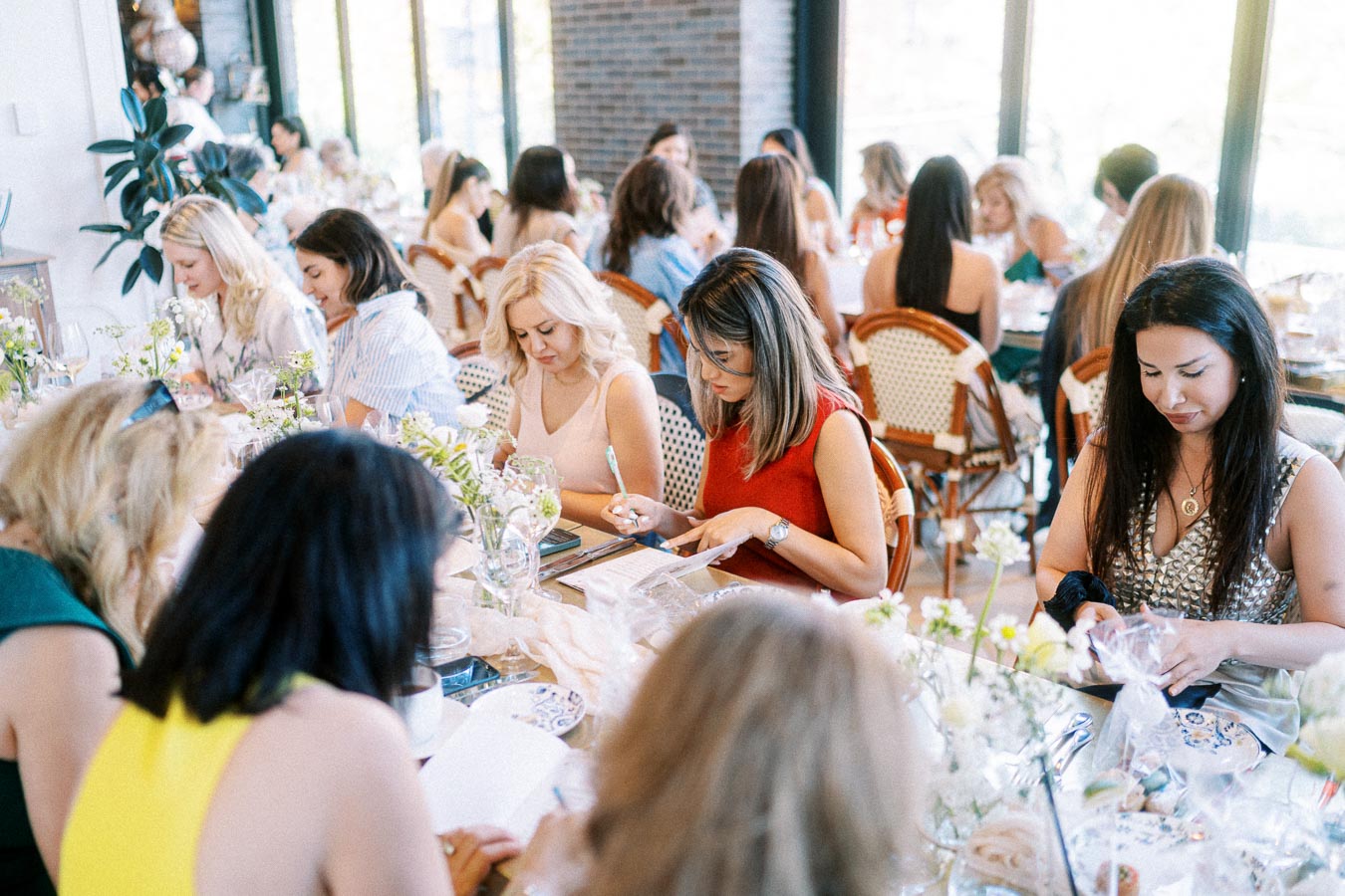 Women enjoying a creative workshop at a stylish cafe, attentively engaging in writing and crafting activities surrounded by floral decor.