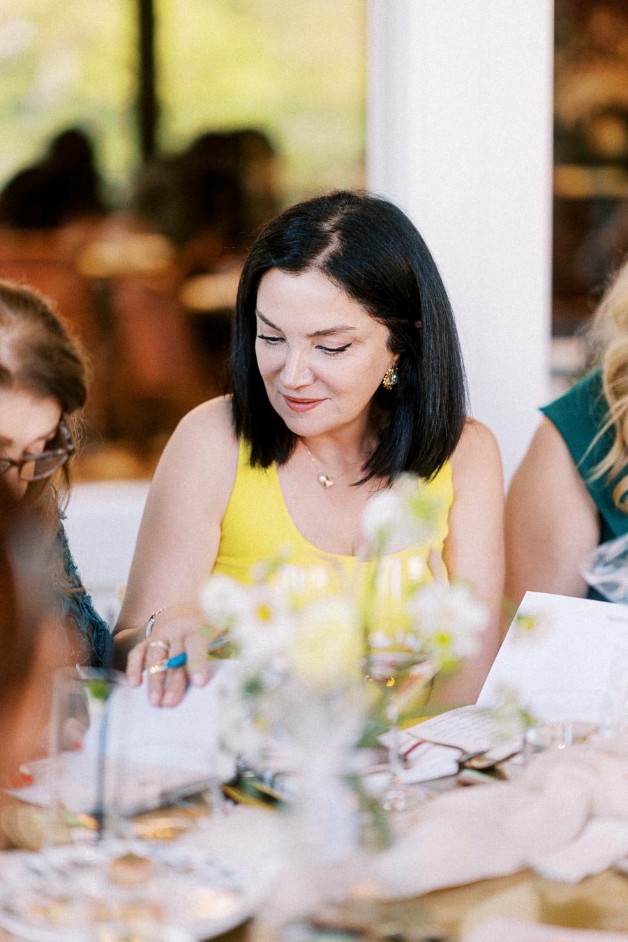 A woman in a yellow dress smiling while sitting at a dining table, surrounded by people, with flowers and tableware in the foreground, creating a warm and inviting atmosphere.