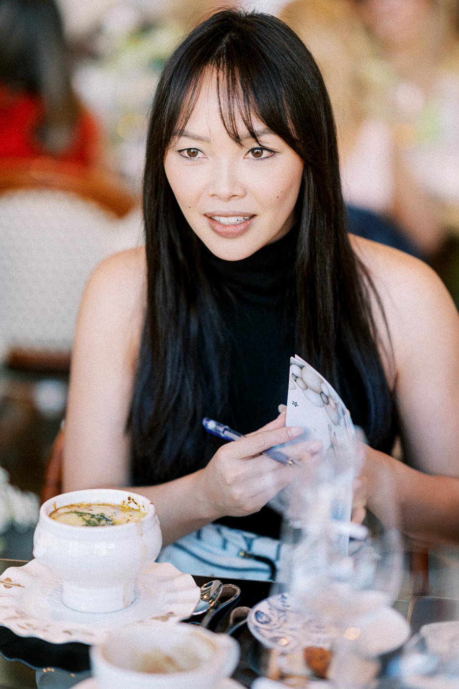 A woman enjoying a meal at a restaurant, holding a pen and paper, with a bowl of soup on the table.